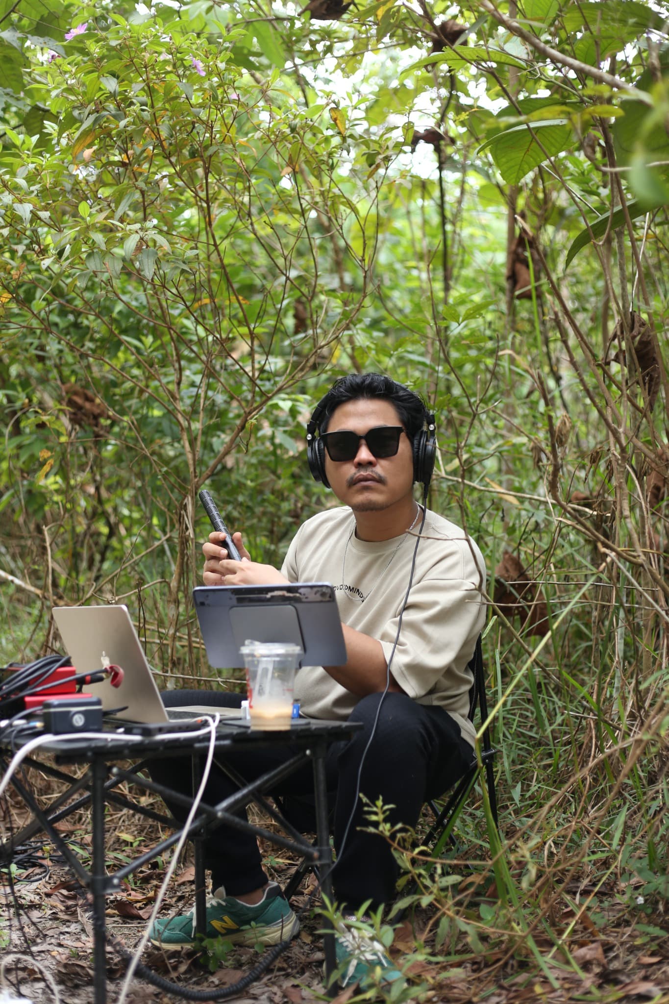Eka di area bervegetasi dengan headphone, mikrofon, dan laptop. Jangan mengarang lokasi atau sesi rekamannya.